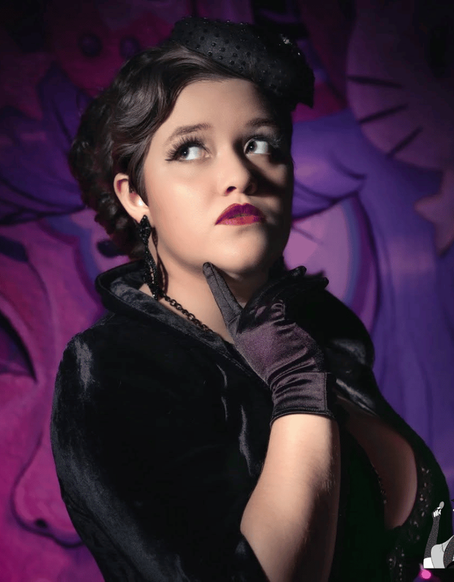 Pale person with makeup dressed in black in front of a purple background