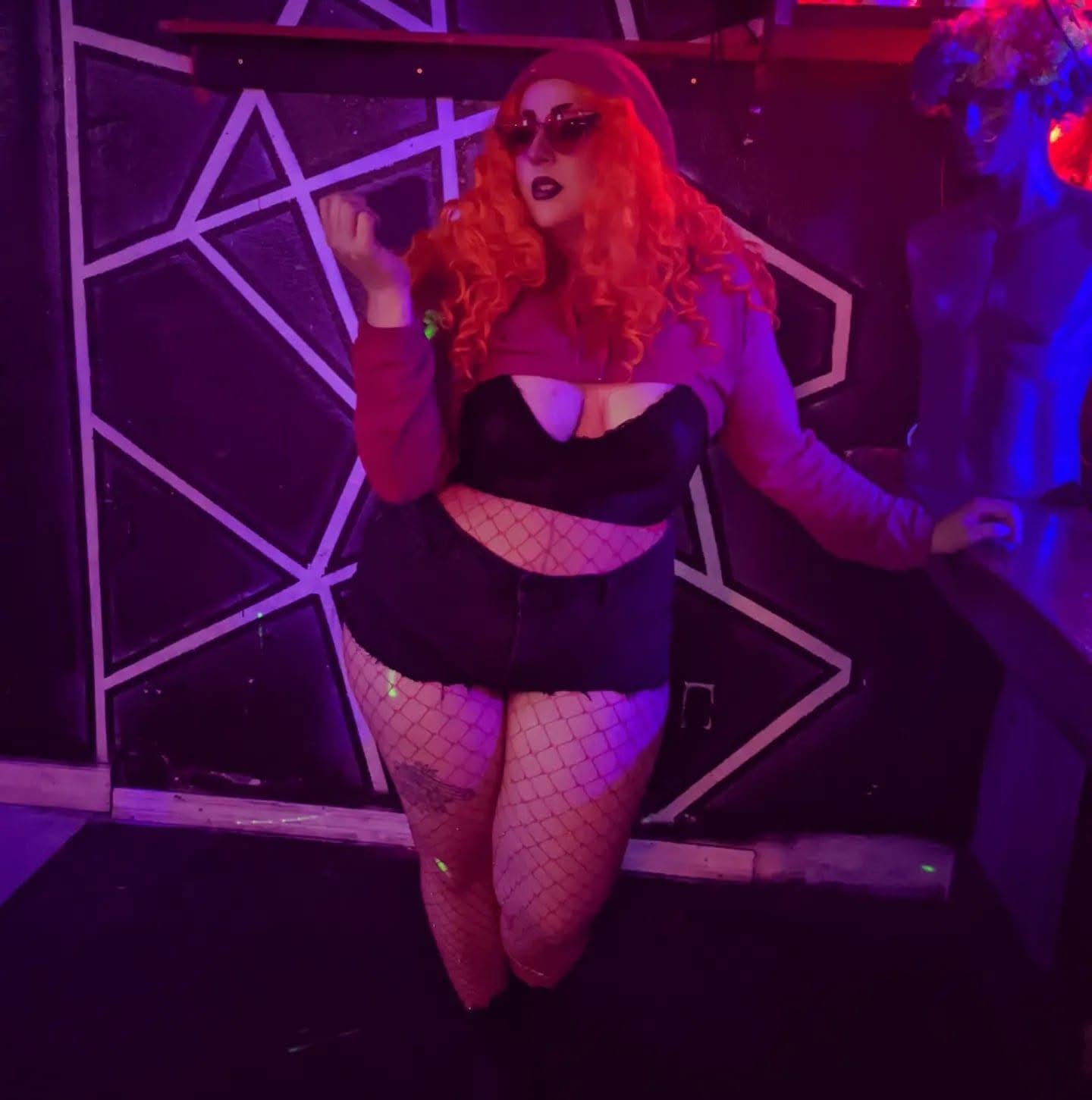 pale person in red curly wig and black outfit in front of geometric pattern
