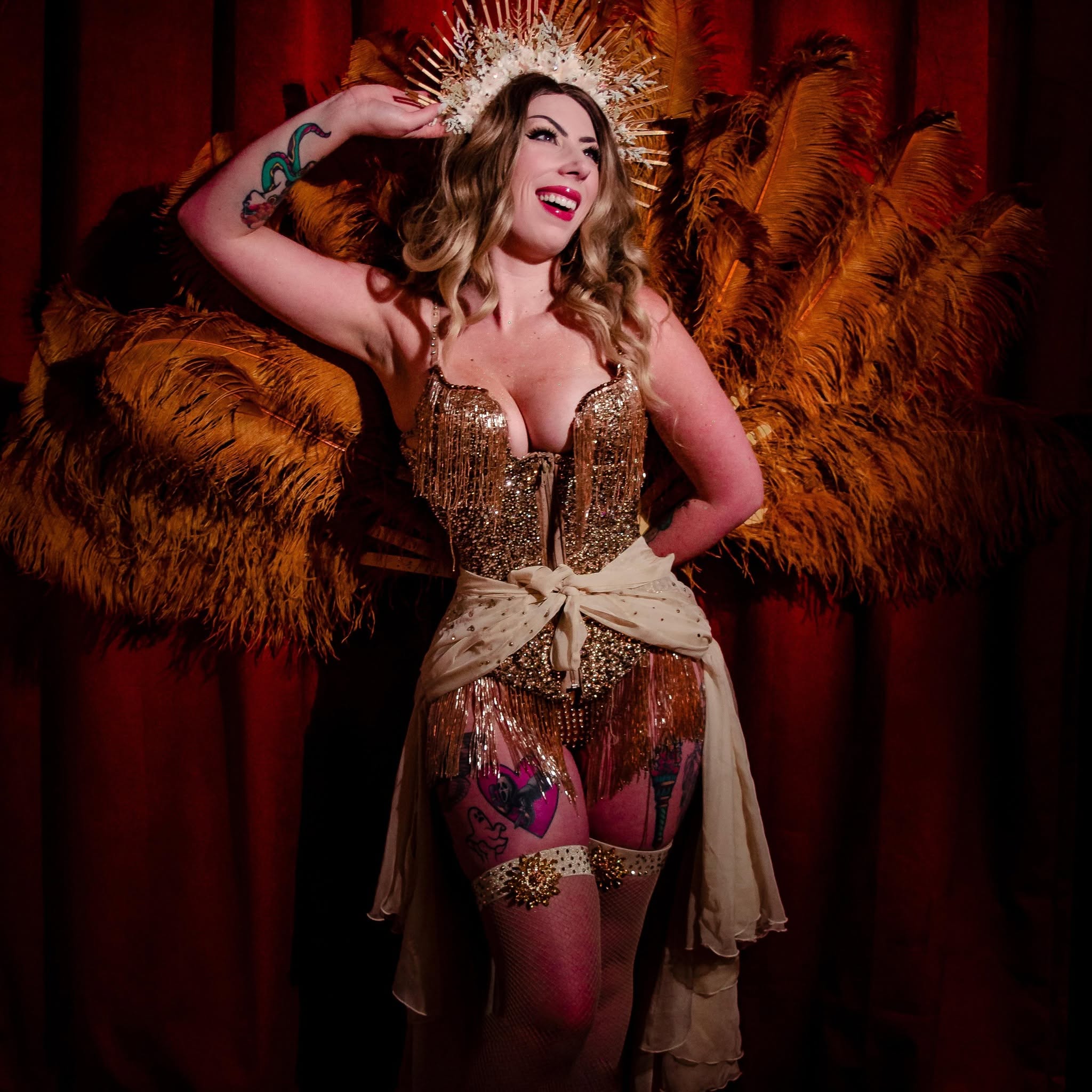 pale burlesque performer in gold ensemble with a gold headpiece standing in front of deep orange feathers